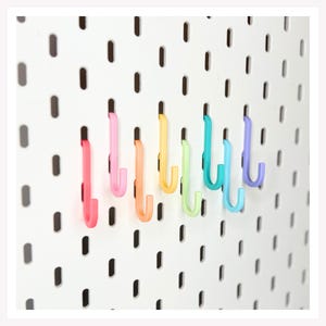 May include: A white pegboard with a series of vertical slots features a row of colorful J-shaped hooks in shades of pink, orange, yellow, light green, turquoise, blue, and purple, each inserted into the board's rectangular openings.
