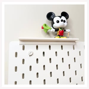 May include: A white pegboard with a matching white shelf attached to the top. A stylized vinyl figure of Mickey Mouse wearing red shorts, yellow shoes, and a green hat is standing on the shelf, holding a small brown tool in one hand.