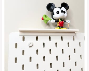May include: A white pegboard with a matching white shelf attached to the top. A stylised vinyl figure of Mickey Mouse wearing red shorts, yellow shoes, and a green hat is standing on the shelf, holding a small brown tool in one hand.