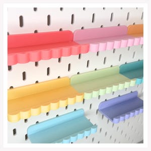 Peut inclure: Une collection d'étagères aux bords festonnés et colorés, fixées sur un panneau perforé blanc. Les étagères sont disposées en rangées et présentent une variété de couleurs pastel et vives, dont le rouge, le rose, l'orange, le jaune, le vert clair, le bleu sarcelle, le bleu et le violet.