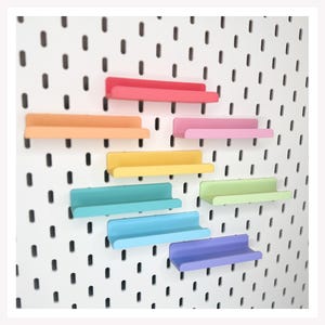 May include: A collection of eight small, horizontal shelves in various colors including red, orange, yellow, light green, teal, light blue, purple, and pink. Each shelf features a raised front edge and is mounted onto a white pegboard panel.