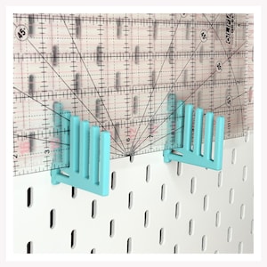 May include: A clear quilting ruler with black grid lines and angle markings is held by two light blue pegboard holders. The holders are attached to a white pegboard. The ruler has metric measurements.