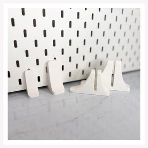 May include: A white pegboard with several white pegboard accessories. The accessories include small rectangular pegs and larger, angled shelf supports. The pegboard has evenly spaced, oval-shaped holes.