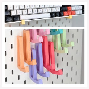 May include: A collection of L-shaped plastic hooks in pastel orange, pink, purple, and green attached to a white pegboard. One yellow hook supports a mechanical keyboard with white, black, and orange keys, including command and option labels.