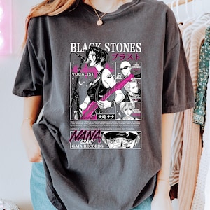 K&ouml;nnte beinhalten: Dunkelgraues T-Shirt mit einer Manga-Illustration einer S&auml;ngerin, die eine rosa Gitarre spielt. Das Shirt enth&auml;lt den Text "BLACK STONES" und japanische Schriftzeichen sowie "NANA OSAKI" und "GAIA RECORDS".