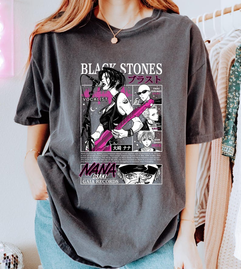 K&ouml;nnte beinhalten: Dunkelgraues T-Shirt mit einer Manga-Illustration einer S&auml;ngerin, die eine rosa Gitarre spielt. Das Shirt enth&auml;lt den Text "BLACK STONES" und japanische Schriftzeichen sowie "NANA OSAKI" und "GAIA RECORDS".