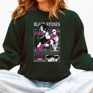 K&ouml;nnte beinhalten: Dunkelgr&uuml;nes Sweatshirt mit einer Manga-Grafik. Die Grafik enth&auml;lt den Text "BLACK STONES" und japanische Schriftzeichen sowie eine Illustration einer Frau, die Gitarre spielt. Das Sweatshirt tr&auml;gt auch den Text "NANA OSAKI GAIA RECORDS".