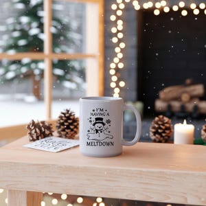 Puede incluir: Una taza de cerámica blanca con el texto "I'M HAVING A MELTDOWN" y un gráfico de muñeco de nieve. La taza está sobre una superficie de madera con piñas y una vela encendida en el fondo. La escena es festiva.