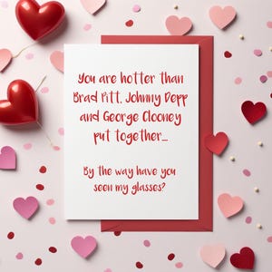 May include: A white card with a red border and red hearts scattered around it. The card reads "You are hotter than Brad Pitt. Johnny Depp and George Clooney put together... By the way have you seen my glasses?"
