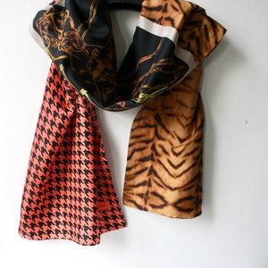 May include: A multi-colored silk scarf with a black, white, and gold pattern on one side and a brown and orange animal print on the other side. The scarf is folded in half and has a pink and black houndstooth pattern on the bottom.