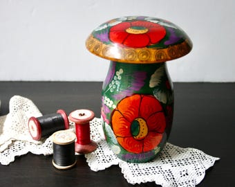 May include: A hand-painted wooden mushroom-shaped container with a green, purple, and red floral design. The container is sitting on a white crocheted doily with spools of thread.