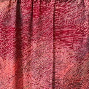 May include: Red and pink fabric curtain with a scribbled, abstract design. The curtain has a gathered top and a flowing, textured appearance. The pattern consists of thin, gray and white lines over a gradient of red and pink hues.