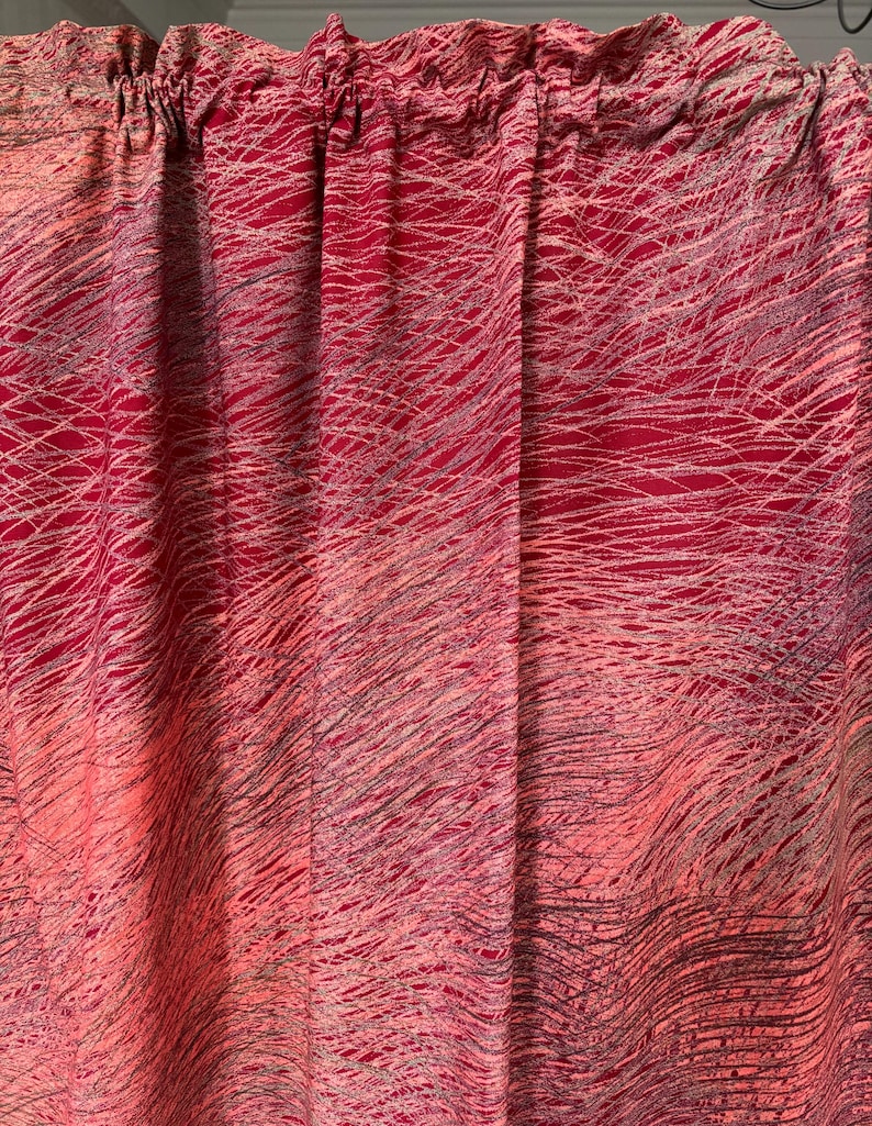 May include: Red and pink fabric curtain with a scribbled, abstract design. The curtain has a gathered top and a flowing, textured appearance. The pattern consists of thin, gray and white lines over a gradient of red and pink hues.
