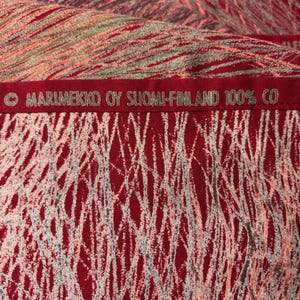May include: Close-up of a red fabric with a white, gray, and pink abstract pattern. The fabric has the text "MARIMEKKO OY SUOMI-FINLAND 100% CO" embroidered in green. The fabric appears to be cotton.