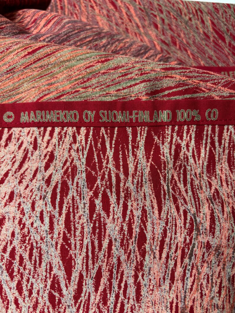 May include: Close-up of a red fabric with a white, gray, and pink abstract pattern. The fabric has the text "MARIMEKKO OY SUOMI-FINLAND 100% CO" embroidered in green. The fabric appears to be cotton.