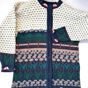 May include: A vintage cardigan sweater with a nature-inspired design. The cream-colored top features a pattern of small black shapes. The body of the sweater has a landscape design with trees, water, and boats. The sweater has a navy blue trim and buttons.