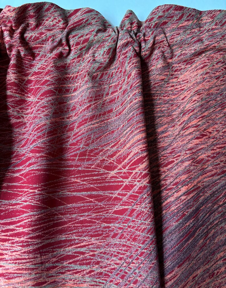 May include: Close-up of a fabric with a red base and a pattern of white and gray lines. The fabric is gathered at the top, creating soft folds. The overall effect is a textured, abstract design.