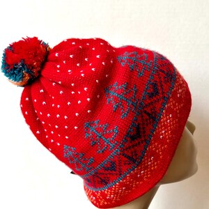 Könnte beinhalten: Eine rote Strickmütze mit einem großen Bommel und einem gemusterten Design. Die Mütze hat weiße Tupfen und türkisfarbene und marineblaue geometrische Muster. Der Bommel hat türkisfarbene, orangefarbene und rote Akzente. Die Mütze ist so konzipiert, dass sie den Kopf warm hält.