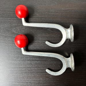 Könnte beinhalten: Zwei Wandhaken aus silberfarbenem Metall, die jeweils mit einem glatten, kugelförmigen roten Knopf am Ende des oberen Arms versehen sind. Die Haken haben einen geschwungenen unteren Teil und eine runde Basisplatte zur Wandmontage.