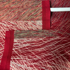 May include: Close-up of red fabric with a white, linear pattern. The fabric has a textured appearance and is accented with a solid red border. The design includes a mix of red, white, and muted green tones, creating a dynamic visual texture.