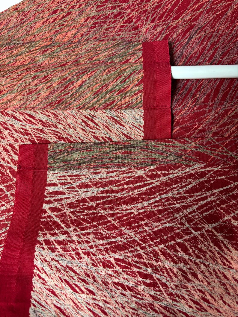May include: Close-up of red fabric with a white, linear pattern. The fabric has a textured appearance and is accented with a solid red border. The design includes a mix of red, white, and muted green tones, creating a dynamic visual texture.