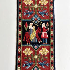 May include: A rectangular, hand-stitched tapestry with a dark blue border. The tapestry features a red background with floral and bird motifs, and a central panel depicting figures in a scene. The tapestry is likely a decorative wall hanging.