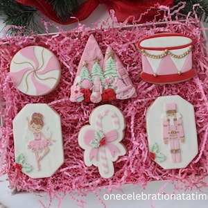 May include: A box of decorated Christmas cookies with a pink and white theme. The cookies include a peppermint swirl, a Christmas tree, a drum, a ballerina, a candy cane, and a nutcracker. The cookies are arranged on pink paper shred.