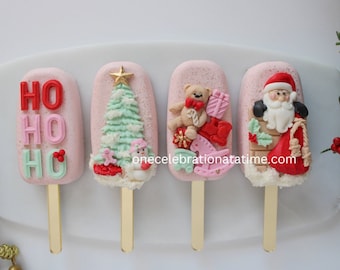 Pode incluir: Quatro picolés cor-de-rosa com diferentes temas natalinos. O primeiro picolé diz "HO HO HO" em letras vermelhas, rosa e verdes. O segundo picolé tem uma árvore de Natal verde com uma estrela dourada no topo. O terceiro picolé tem um urso de pelúcia segurando um bastão de caramelo e um presente. O quarto picolé tem o Papai Noel descendo pela chaminé com um saco de presentes.