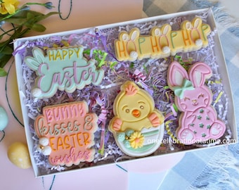 Pode incluir: Uma caixa de presente cheia de biscoitos de Páscoa em tons pastel. Os biscoitos são decorados com desenhos de coelhos, pintinhos e ovos. A caixa está cheia de papel picado e os biscoitos estão dispostos em um padrão decorativo. Os biscoitos têm várias mensagens com tema de Páscoa, incluindo "Feliz Páscoa", "Beijos de coelho e desejos de Páscoa" e "Hop Hop Hop".