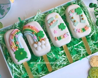 Pode incluir: Quatro cake pops cobertos de chocolate branco decorados com temas do Dia de São Patrício. Os pops estão dispostos em uma caixa branca forrada com papel verde picado. Os pops são decorados com arco-íris, trevos, um pote de ouro, um chapéu de duende, uma ferradura e as palavras "Lucky" e "Charms".