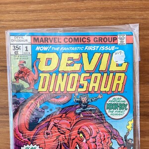 May include: A comic book cover featuring a red dinosaur with sharp teeth and claws. The dinosaur is standing on a rocky surface with smaller figures in the background. The title of the comic is "Devil Dinosaur" and the issue number is 1. The text on the cover also says "Now! The Fantastic First Issue!" and "Also Featuring: (Moon-Boy) The First Human!"