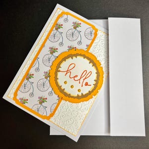 May include: A handmade greeting card featuring a white base with an embossed floral pattern, a panel with a repeating bicycle and flower basket print, and a circular orange scalloped die-cut topper with the handwritten orange text hello.