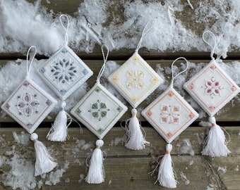 Op de afbeelding: Een verzameling van zeven ruitvormige, geborduurde ornamenten met geometrische patronen in pasteltinten. Elk ornament heeft een witte kwast en een lus om op te hangen. De ornamenten liggen op een houten oppervlak met sneeuw.
