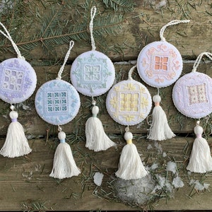 Può includere: Sei ornamenti ricamati bianchi con nappe. Gli ornamenti sono rotondi e presentano diversi disegni di ricamo colorati. Gli ornamenti sono appesi a una superficie di legno.