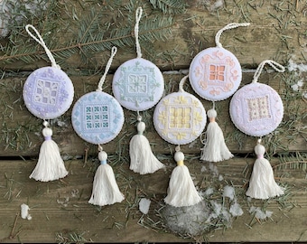 Op de afbeelding: Zes witte geborduurde ornamenten met kwastjes. De ornamenten zijn rond en hebben verschillende gekleurde borduurmotieven. De ornamenten hangen aan een houten oppervlak.