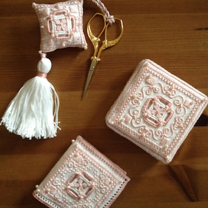 Peut inclure: Deux boîtes en tissu brodées de rose et de blanc avec un pompon blanc et des ciseaux dorés. Les boîtes sont carrées et présentent un motif floral délicat.