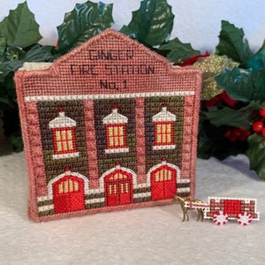 May include: A hand-stitched miniature fire station with the words "Ginger Fire Station No. 1" on the front. The fire station is red, green, and white with three windows and three garage doors. A small red and white wagon with a horse is in the foreground.