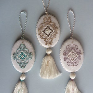 May include: Three oval-shaped ornaments with embroidered designs and white tassels. The ornaments are white with different colored embroidery: gold, teal, and pink.