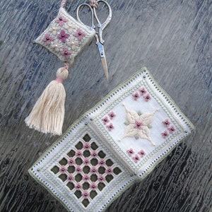 Peut inclure: Un porte-ciseaux en point de croix blanc et vert avec un motif floral rose. Le porte-ciseaux est plié en deux et possède un petit coussin à aiguilles floral blanc et rose qui y est attaché avec un pompon.