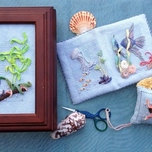 Puede incluir: Un diseño de punto de cruz enmarcado que presenta un caballito de mar azul, algas verdes y una rama. También hay varias otras piezas de punto de cruz, incluyendo una almohada, un libro pequeño y una estrella de mar.