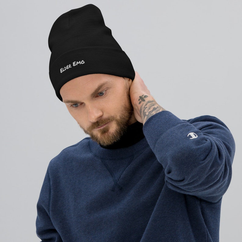 May include: A black beanie with the text "ELDER EMO" embroidered in white on the front. The beanie is being worn by a person with a beard and a tattoo on their arm.