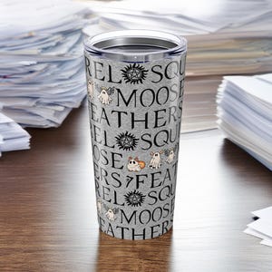 May include: A stainless steel tumbler with a silver lid. The tumbler has a gray, textured background with the words "Squirrel, Moose, Feathers" and other text. The design includes cartoon ghosts, a dog, and a sun symbol.