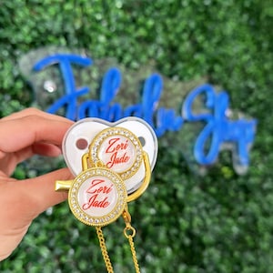 May include: A white baby pacifier with a gold-colored clip and chain. The pacifier features two round, decorative elements with the name "Zori Jade" in red script, surrounded by a ring of small, clear rhinestones. The background is a green, leafy texture.