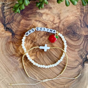 May include: A white pearl bracelet with a gold chain and a charm with the words "MRS CELINA" and a red apple.