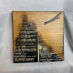 May include: A wooden sign with a burnt edge, featuring a list of bands and events. The text includes "Rock Bowling: The Casualties", "Rock Bowling: Face to Face", "Rock Bowling: A Global Threat", "The Rocket Summer", and "303.777.3278 550 Grant St. Denver".