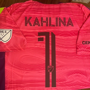 May include: A pink and purple soccer jersey with the name "KAHLINA" and the number "11" printed on the back in black. The MLS logo is on the left sleeve, and the Centene Corporation logo is on the right sleeve.