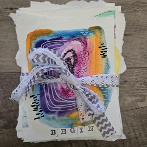 May include: A stack of rectangular paper sheets with deckled edges, tied together with a white lace and grey chevron ribbon. The top sheet features an abstract watercolor painting in orange, purple, and blue with the word BEGIN printed at the bottom.
