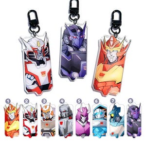 May include: A set of eight acrylic keyrings featuring illustrations of Transformers characters. Each keyring has a black metal clasp. The characters are in various colours, including red, orange, blue, purple, and grey.