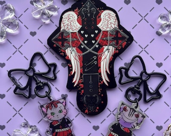 May include: Black and red cross charm with angel wings, safety pins, and chains. The charm is attached to a black metal key chain with two black bow charms. Two small, clear acrylic charms of cats in black and red outfits are also attached to the key chain.