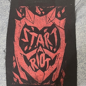 Op de afbeelding: Zwarte stoffen patch met een rood grafisch ontwerp. Het ontwerp bevat de woorden "START RIOT" in een gestileerd lettertype in een gezichtsvorm. De patch is ongeveer 10 cm breed.